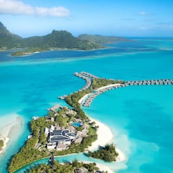 Overwater bungalows connect to turquoise waters via a curving walkway, surrounded by lush greenery on a secluded island.