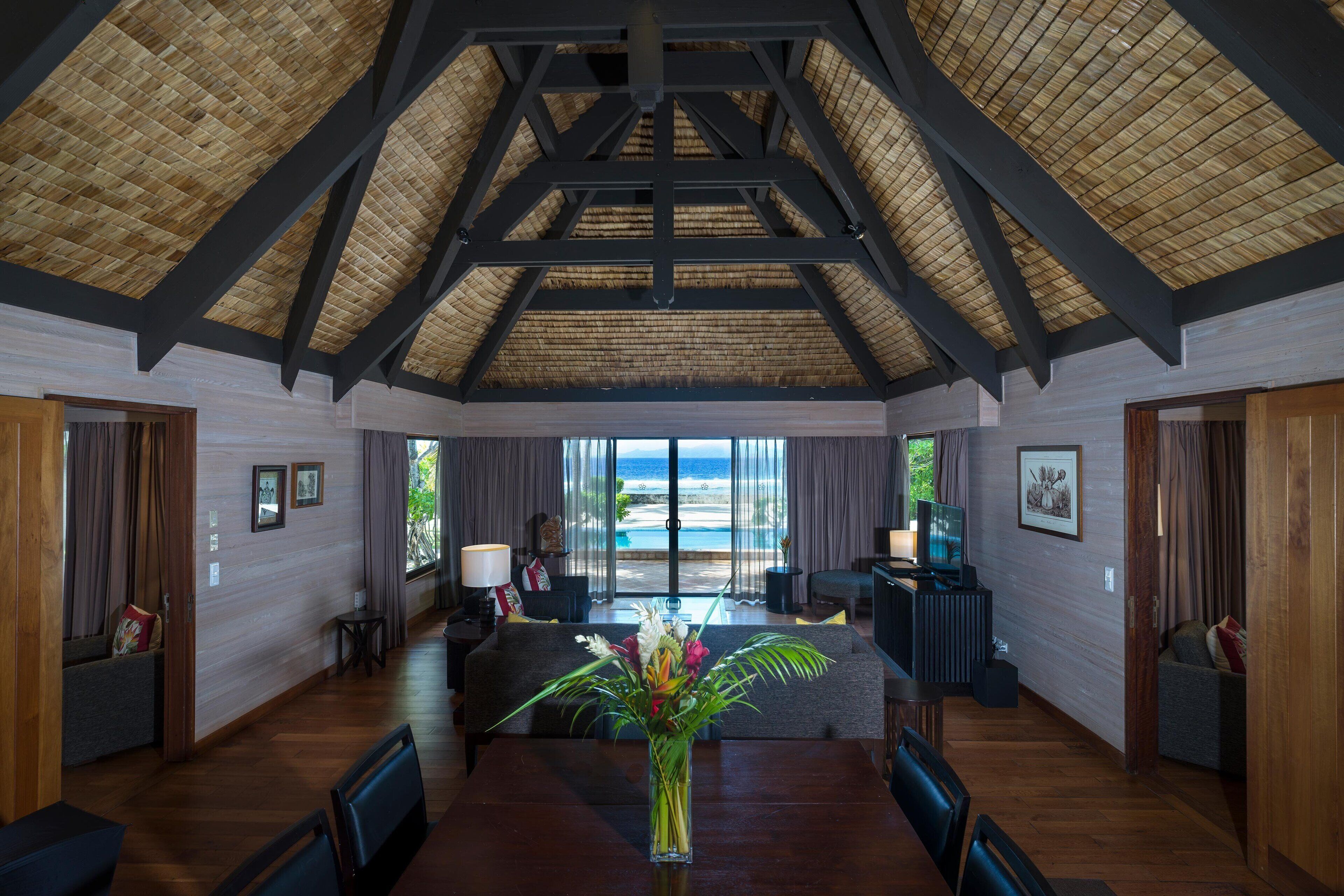 A spacious room with a high ceiling, wooden beams, a large window with a view of the ocean, a dining table with chairs, and a seating area with a sofa and a TV.