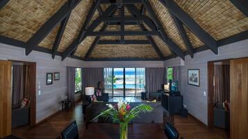 A spacious room with a high ceiling, wooden beams, a large window with a view of the ocean, a dining table with chairs, and a seating area with a sofa and a TV.