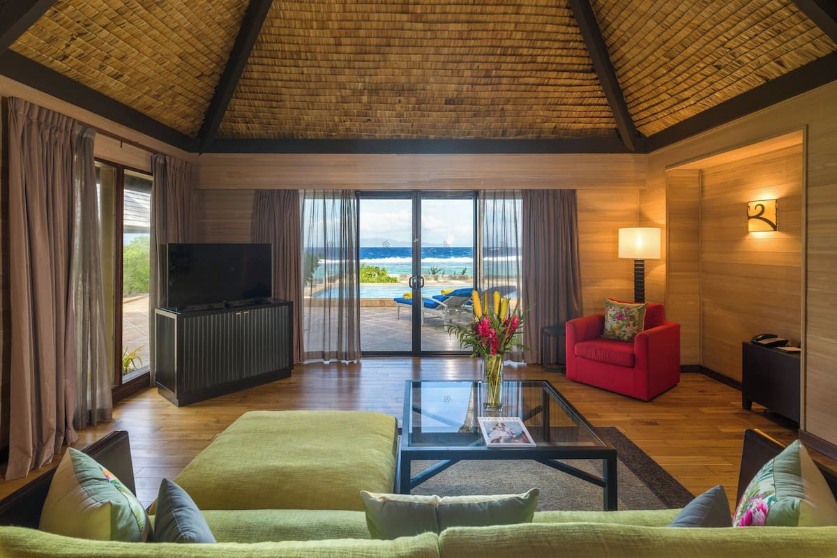 Uma sala de estar ampla com uma televisão grande, assentos confortáveis, uma mesa de centro de vidro e vista para o oceano através de grandes janelas.