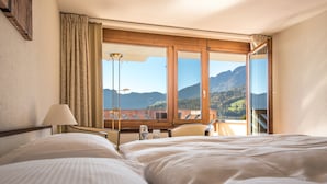 Habitación de hotel con dos camas, un escritorio, una silla y un amplio ventanal con vistas a la montaña.