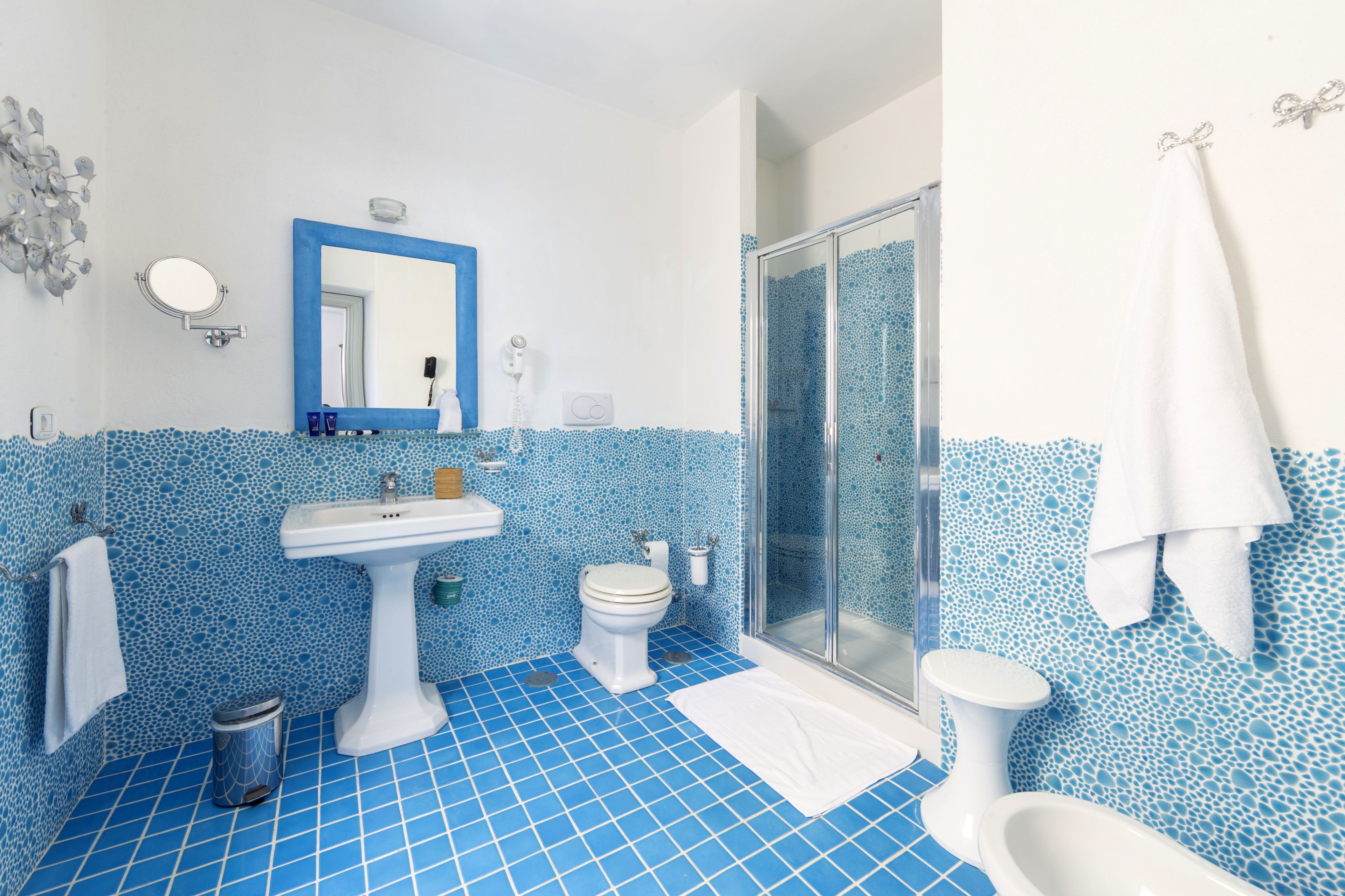 Une salle de bain avec un sol en mosaïque bleue, un lavabo sur pied, deux toilettes, une douche en verre et une serviette blanche accrochée au mur.