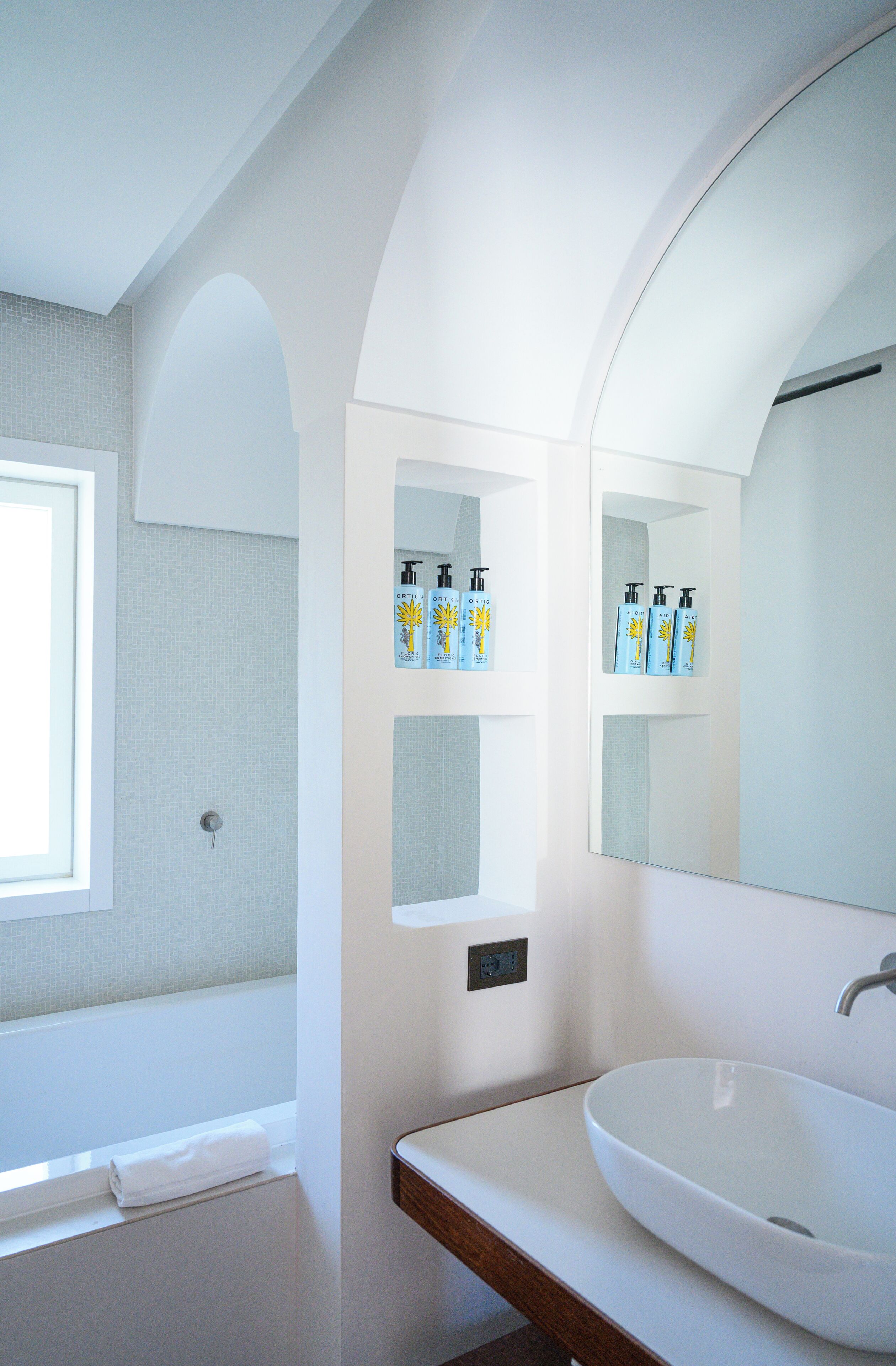Ein Badezimmer mit einem weißen Waschbecken, einem großen Spiegel und Regalen mit Toilettenartikeln.