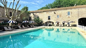 Zona de piscina con sillas de descanso y sombrillas, edificios de piedra y cielo despejado.