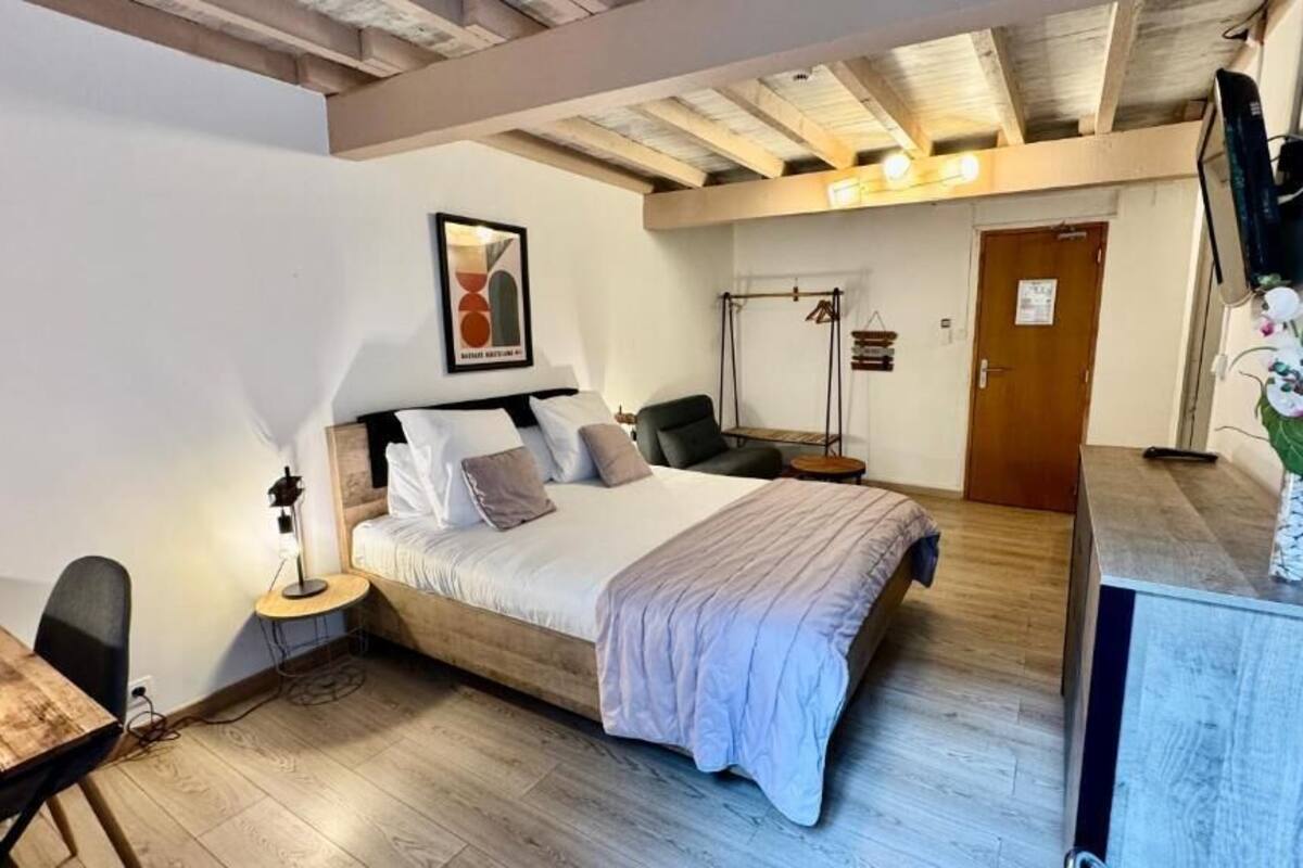A bedroom with a bed, bedside table, chair, and a desk. There is a door and a wall-mounted TV.