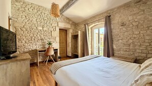 Un dormitorio con pared de piedra, una cama, un escritorio con silla, un televisor y una ventana con cortinas.