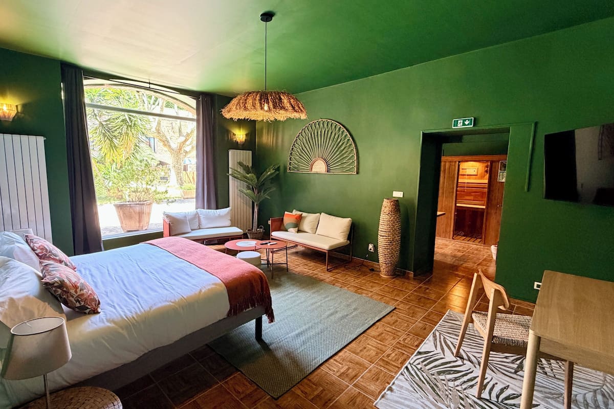 A room with a bed, a sofa, a small table, a lamp, a hanging light fixture, a potted plant, and a view of the outdoors.