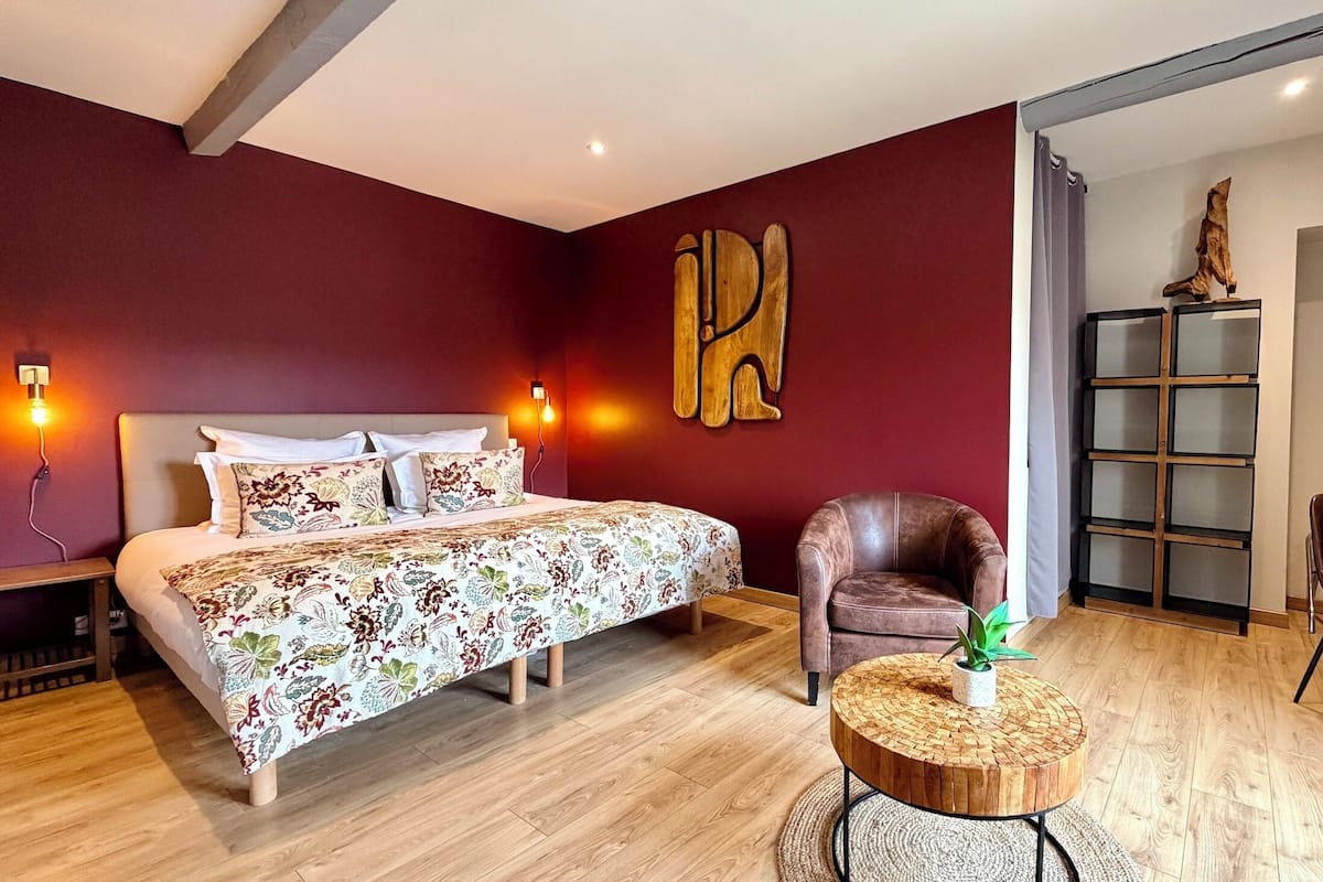 A bedroom with a bed, a chair, a round wooden table, and a bookshelf.