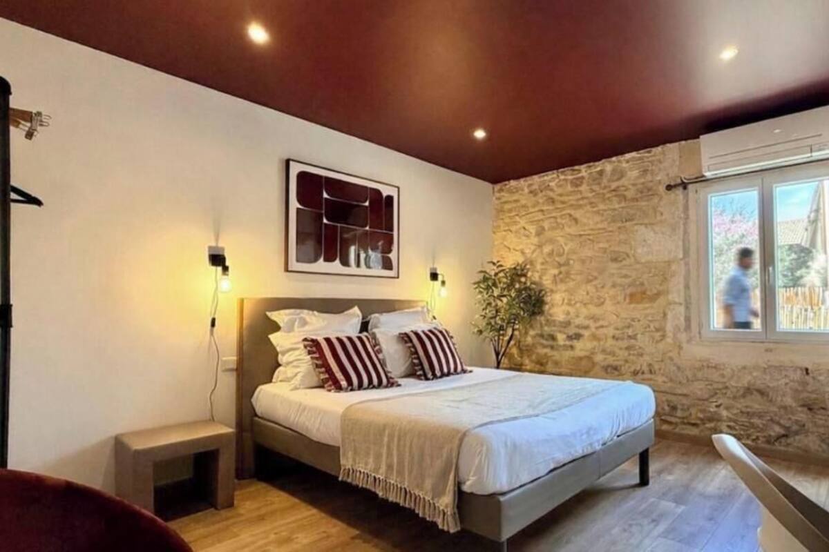 A bedroom with a stone wall, a bed with white bedding, a nightstand, a wall-mounted light, and a window with a view.