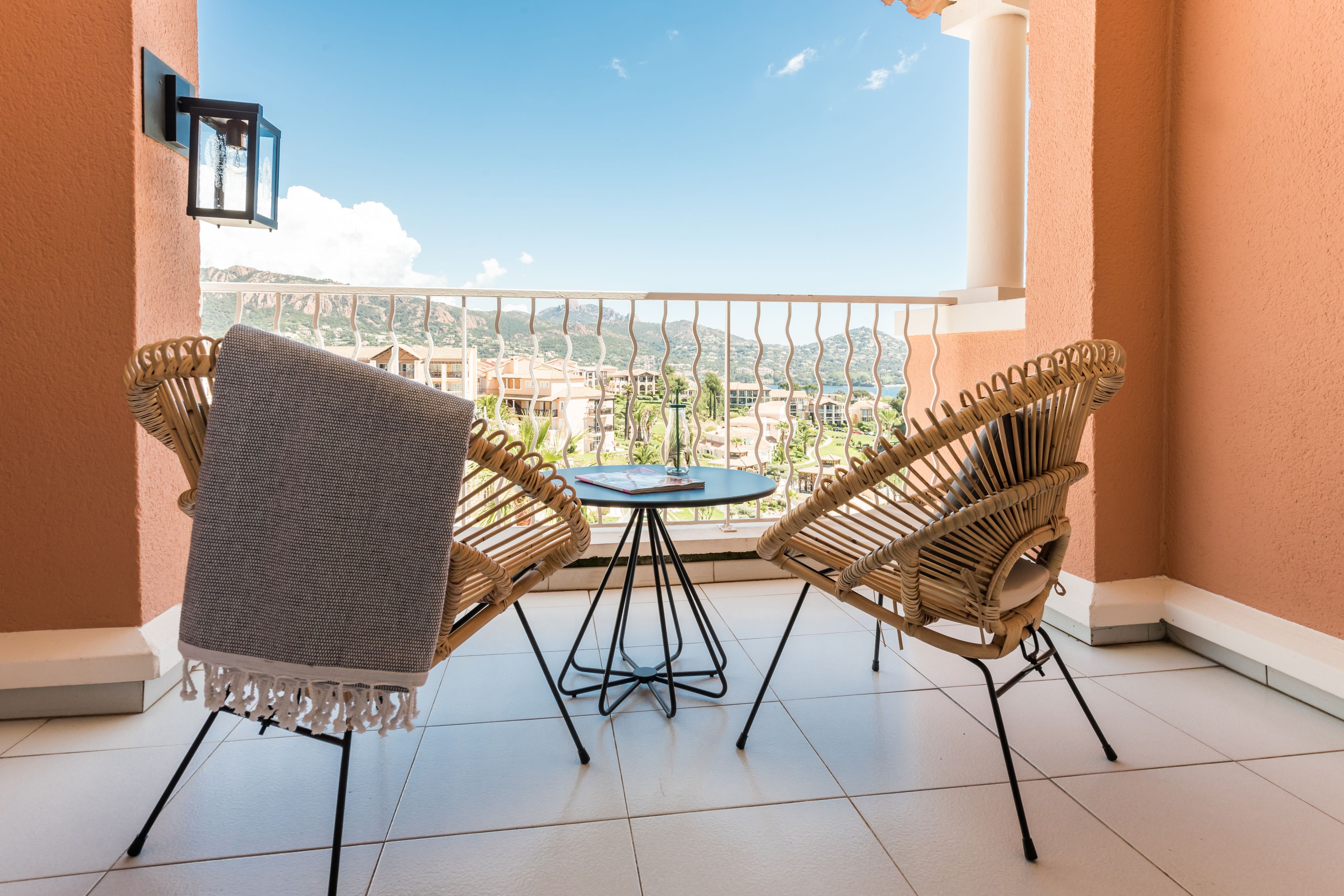 Een balkon met twee rieten stoelen en een kleine ronde tafel, met uitzicht op een schilderachtig stadsbeeld en glooiende heuvels.
