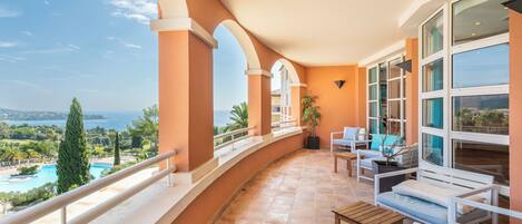 A balcony with a view of a pool and landscape, furnished with white chairs and a wooden table.