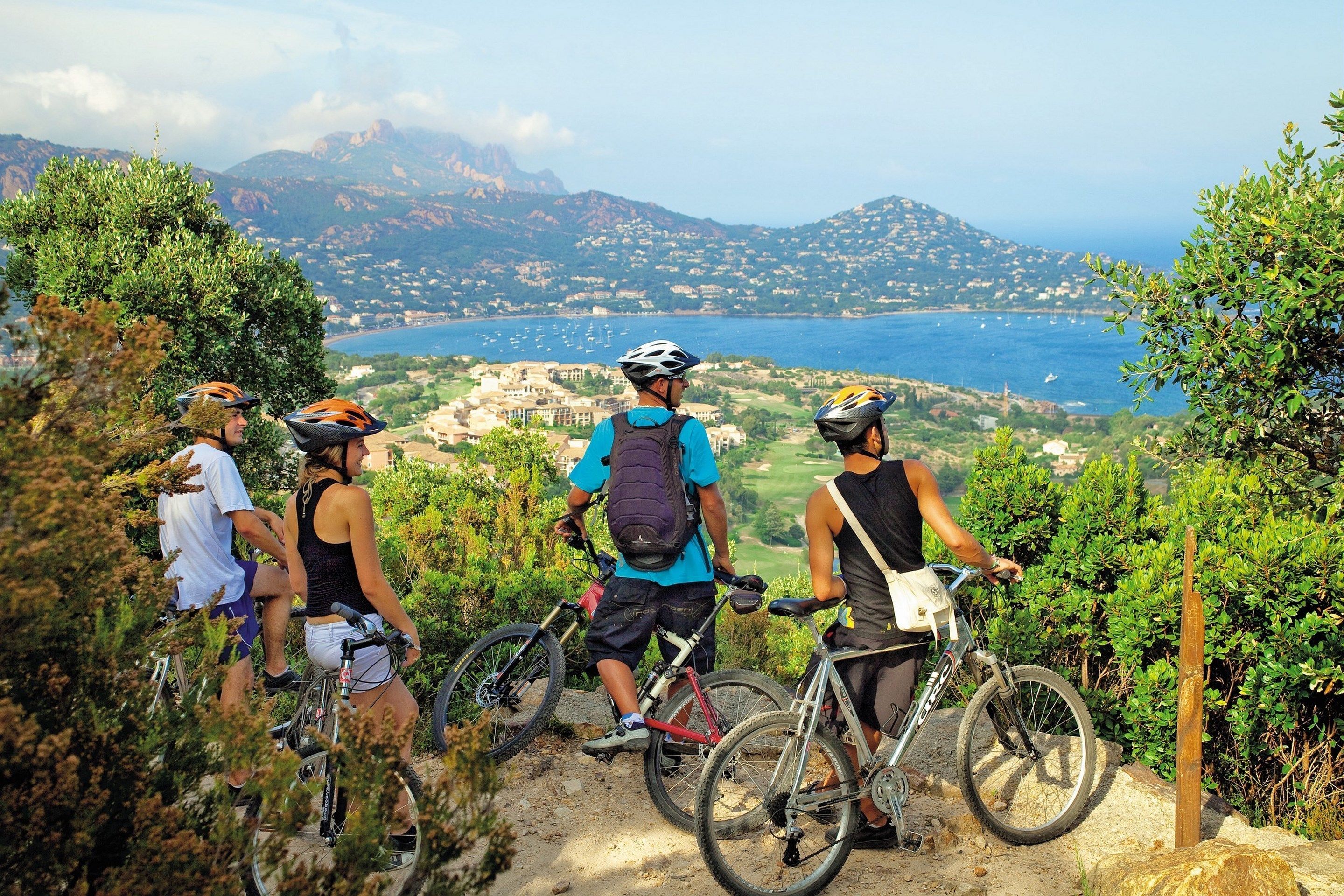 A group of cyclists on a mountain path with a scenic view of a coastal town and mountains in the background.