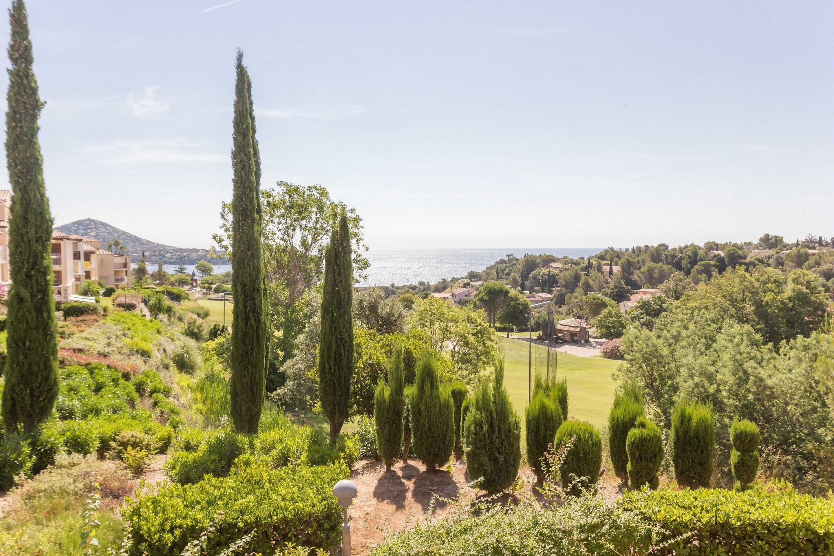 A landscaped garden with tall cypress trees, a pathway, and a view of a coastal town in the background.