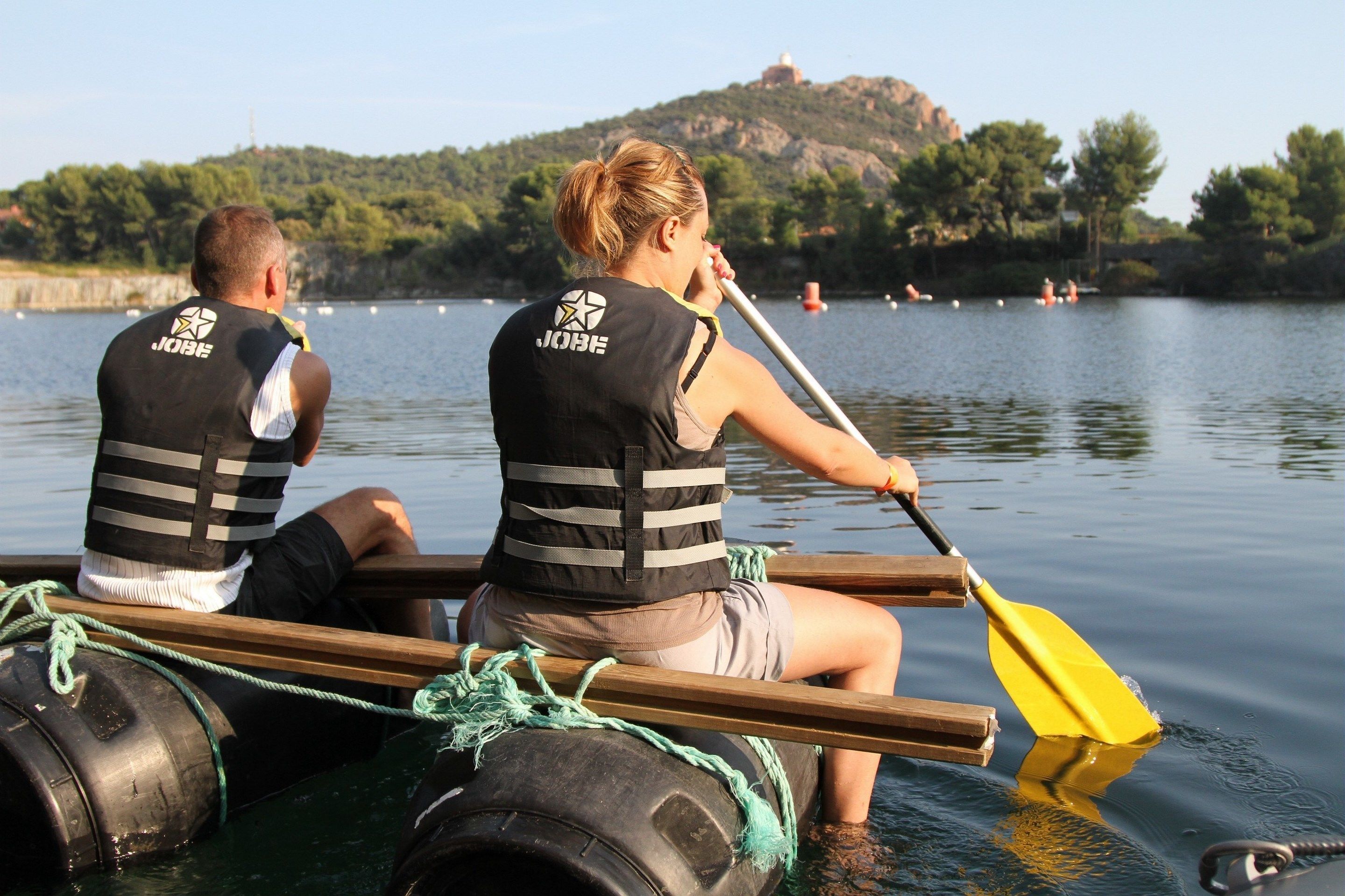 Deux personnes dans un bateau, portant des gilets de sauvetage, pagayant sur une eau calme, avec un paysage vallonné en arrière-plan.