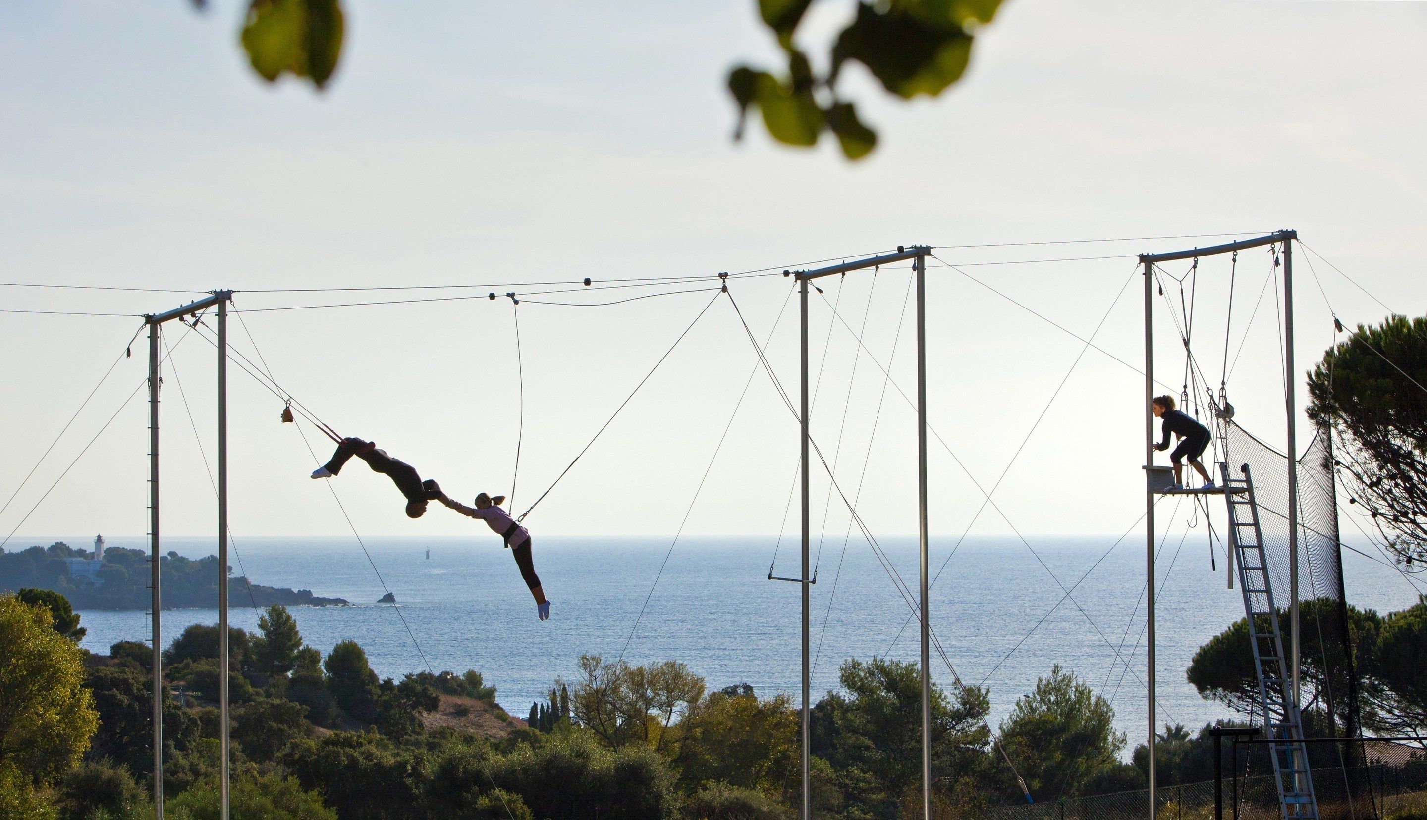 Une installation de trapèze avec une personne qui se balance, une structure en filet et une vue sur la mer et les bâtiments éloignés.