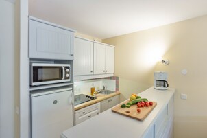 A compact kitchen with white cabinets, a microwave, a coffee maker, and a wooden cutting board with vegetables.