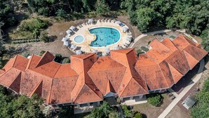 Une vue aérienne d’un complexe hôtelier doté d’une piscine entourée de chaises longues et de parasols, ainsi que d’un bâtiment dont le toit est recouvert de tuiles rouges.