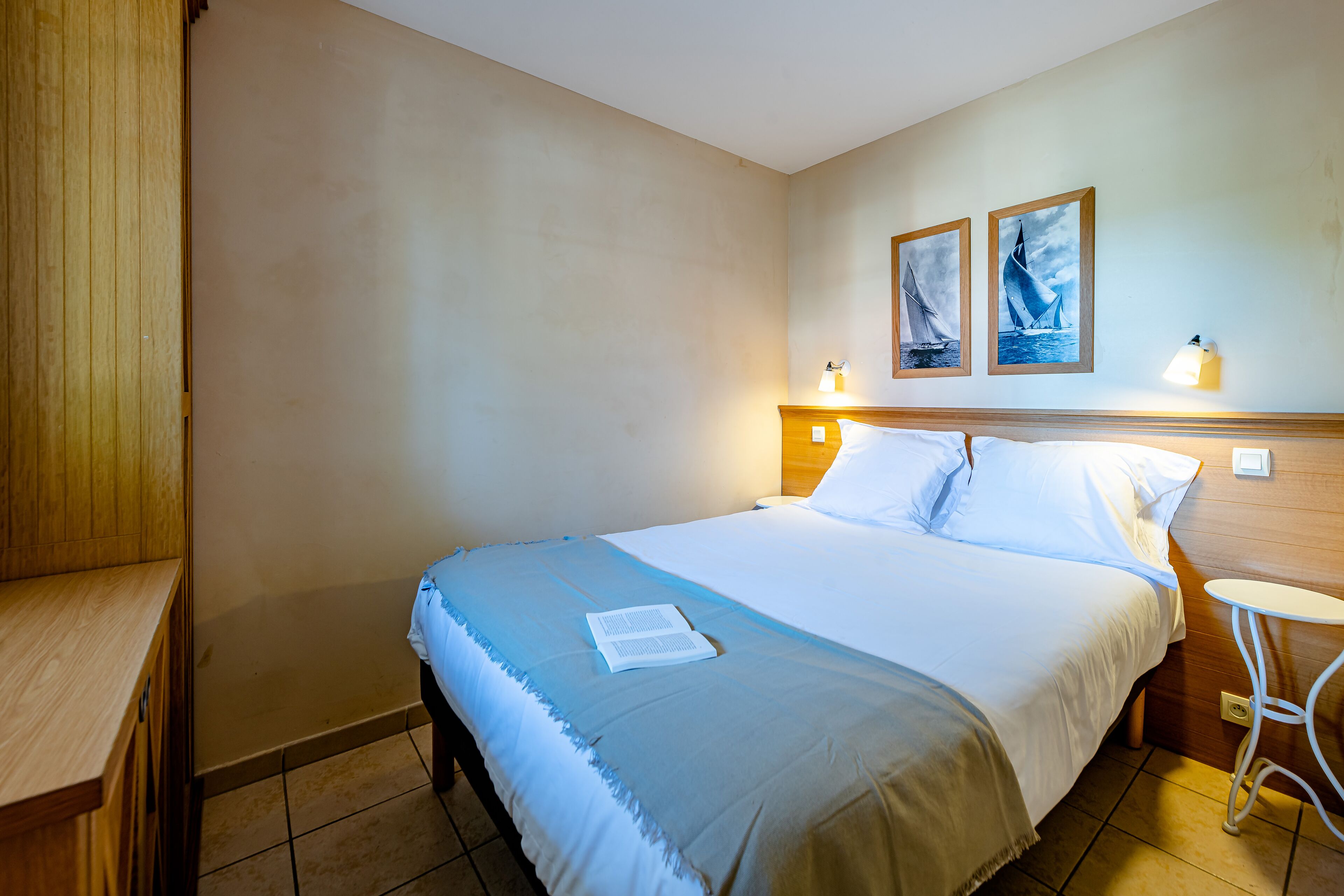 Ein ordentlich bezogenes Bett mit einer blau-weißen Tagesdecke, ein Nachttisch mit Lampe und zwei gerahmte Bilder an der Wand.
