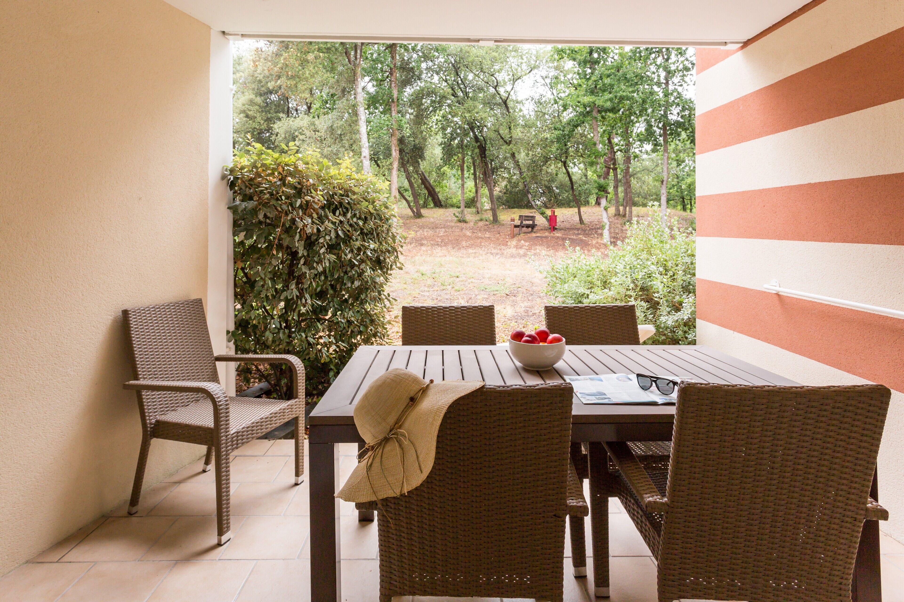 A patio with wicker furniture, a table with a bowl of fruit, and a view of a forested area.