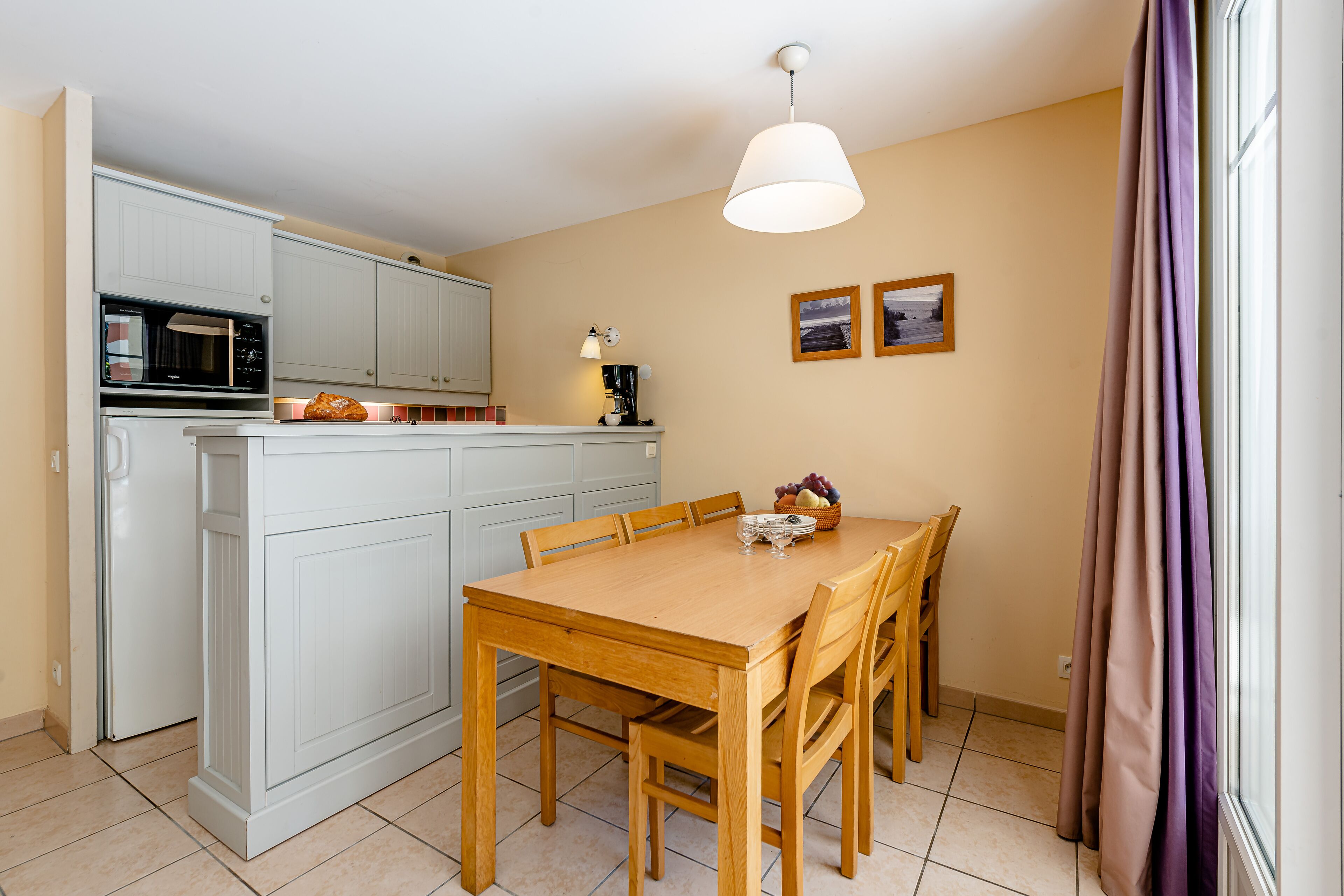 A compact kitchen with white cabinets, a microwave, and a coffee machine. There is a dining table with chairs and a fruit bowl.