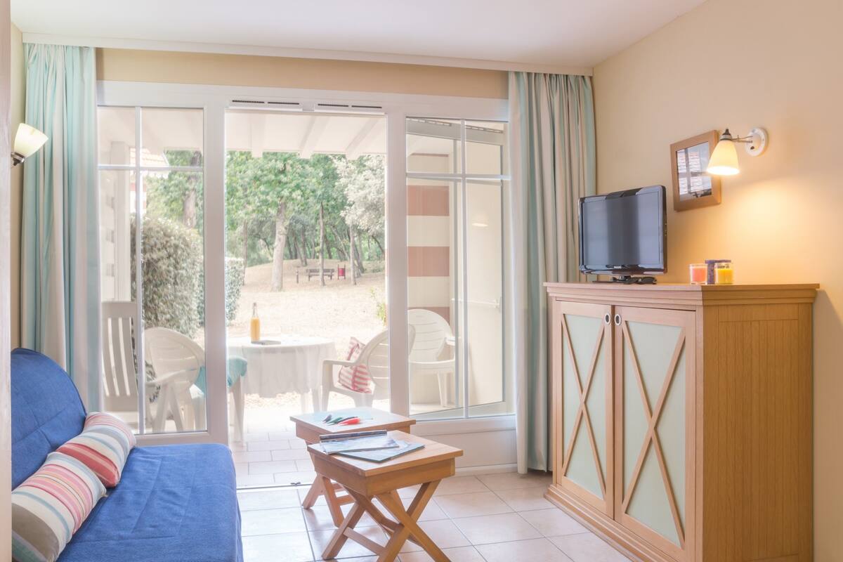 Ein Zimmer mit einem Bett, einem Schreibtisch, einem Fernseher und einer Glastür, die zu einem Außenbereich mit Stühlen und einem Tisch führt.