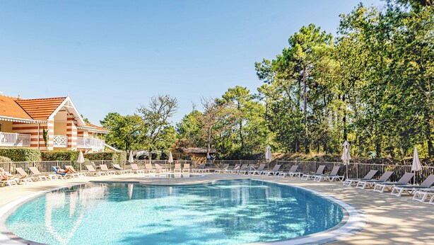 Une piscine entourée d’arbres et d’un bâtiment à toit de tuiles, avec des chaises longues et des parasols à proximité.