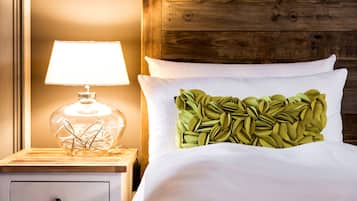 Un lit bien fait, avec du linge de lit blanc et un coussin orné d’un motif de feuilles vertes. Une lampe en verre transparent, dont le fil est enroulé à l’intérieur, est posée sur une table de chevet en bois, à côté du lit.
