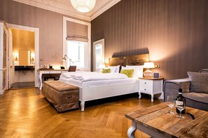 A hotel room with a large bed, a wooden coffee table with a wine bottle and glass, a nightstand with a lamp, a sofa, and a desk with a lamp and a mirror. - Hotel Alsterblick (Hamburg)