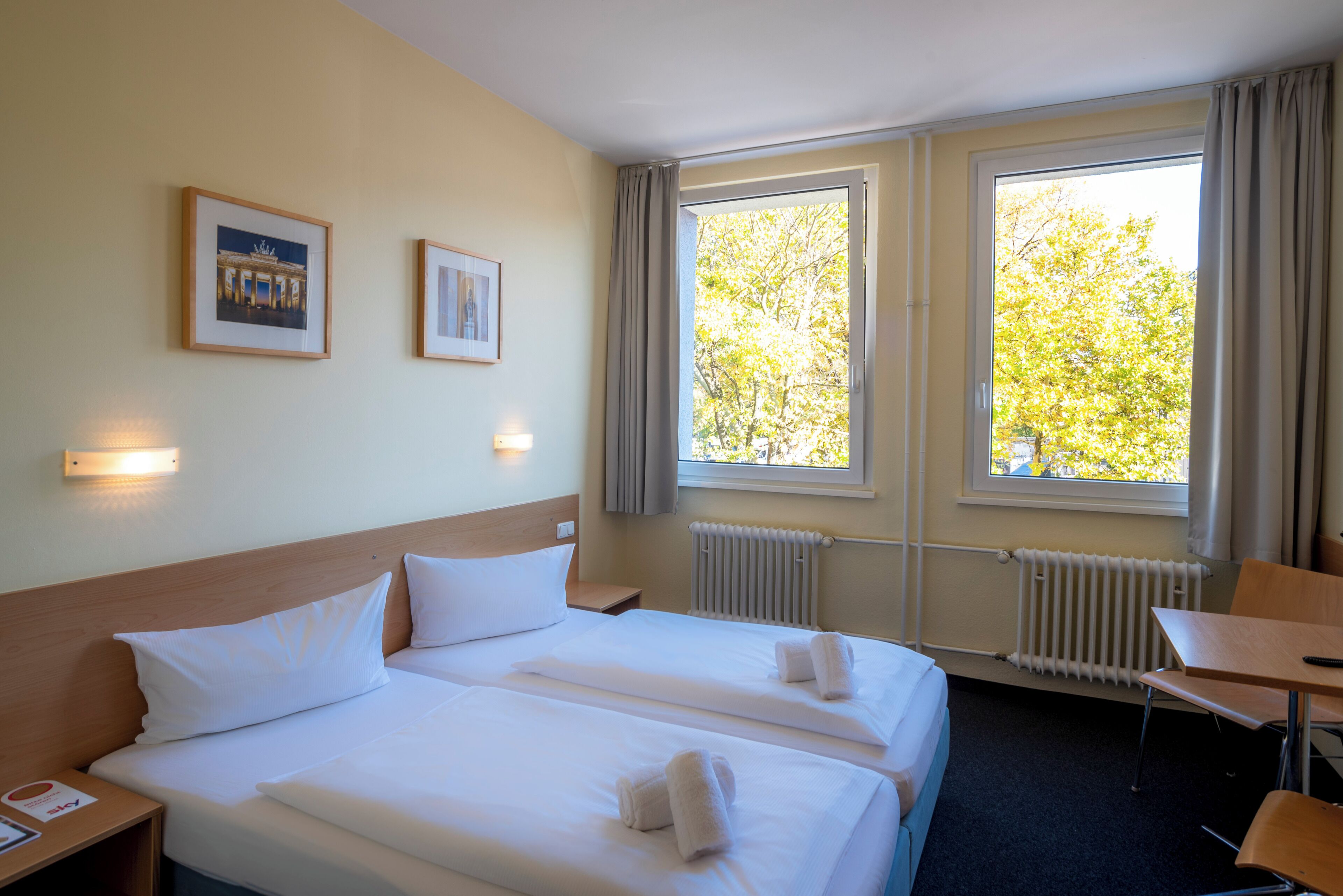 A hotel room with two beds, a desk, a chair, a radiator, and two framed pictures on the wall.
