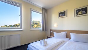 Een hotelkamer met twee bedden, een groot raam, een radiator en twee ingelijste foto's aan de muur.