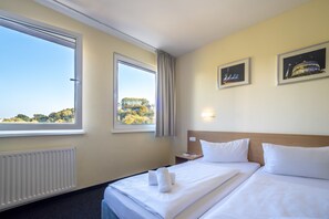Habitación de hotel con dos camas, un ventanal grande, un radiador y dos cuadros en la pared.