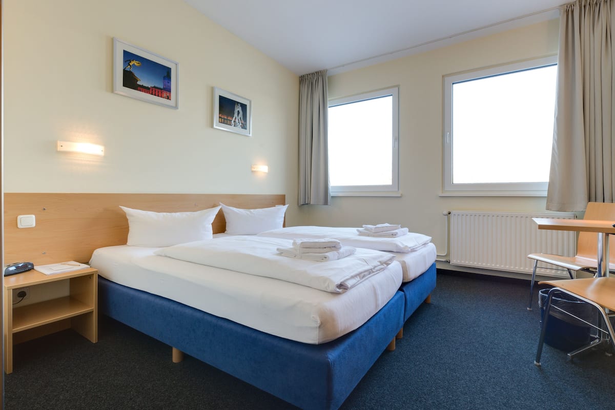 Een hotelkamer met een tweepersoonsbed, een bureau, een stoel, een nachtkastje en twee ramen.