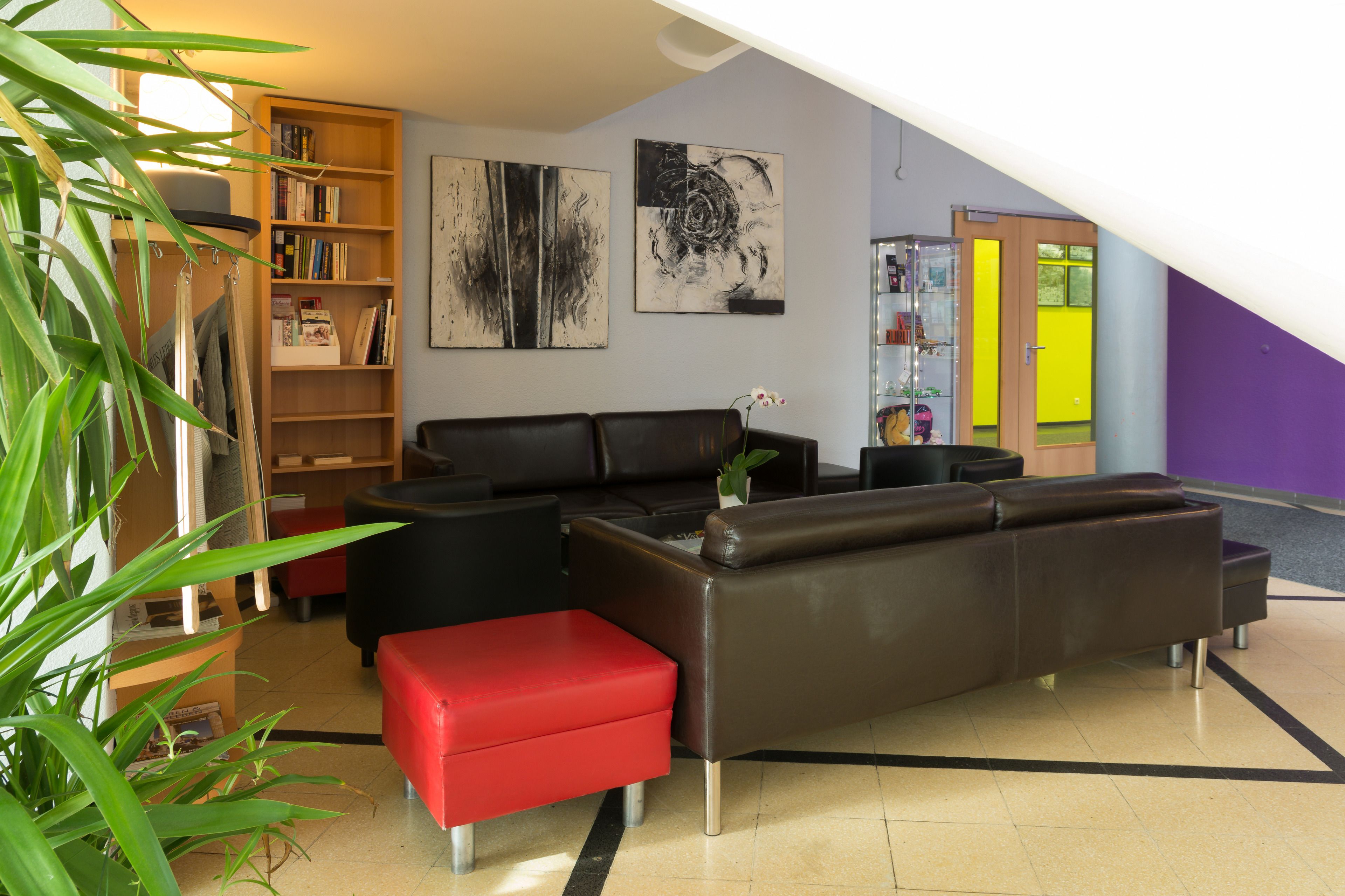 A modern living room with a brown leather sofa, a red ottoman, a bookshelf, and two abstract paintings on the wall.