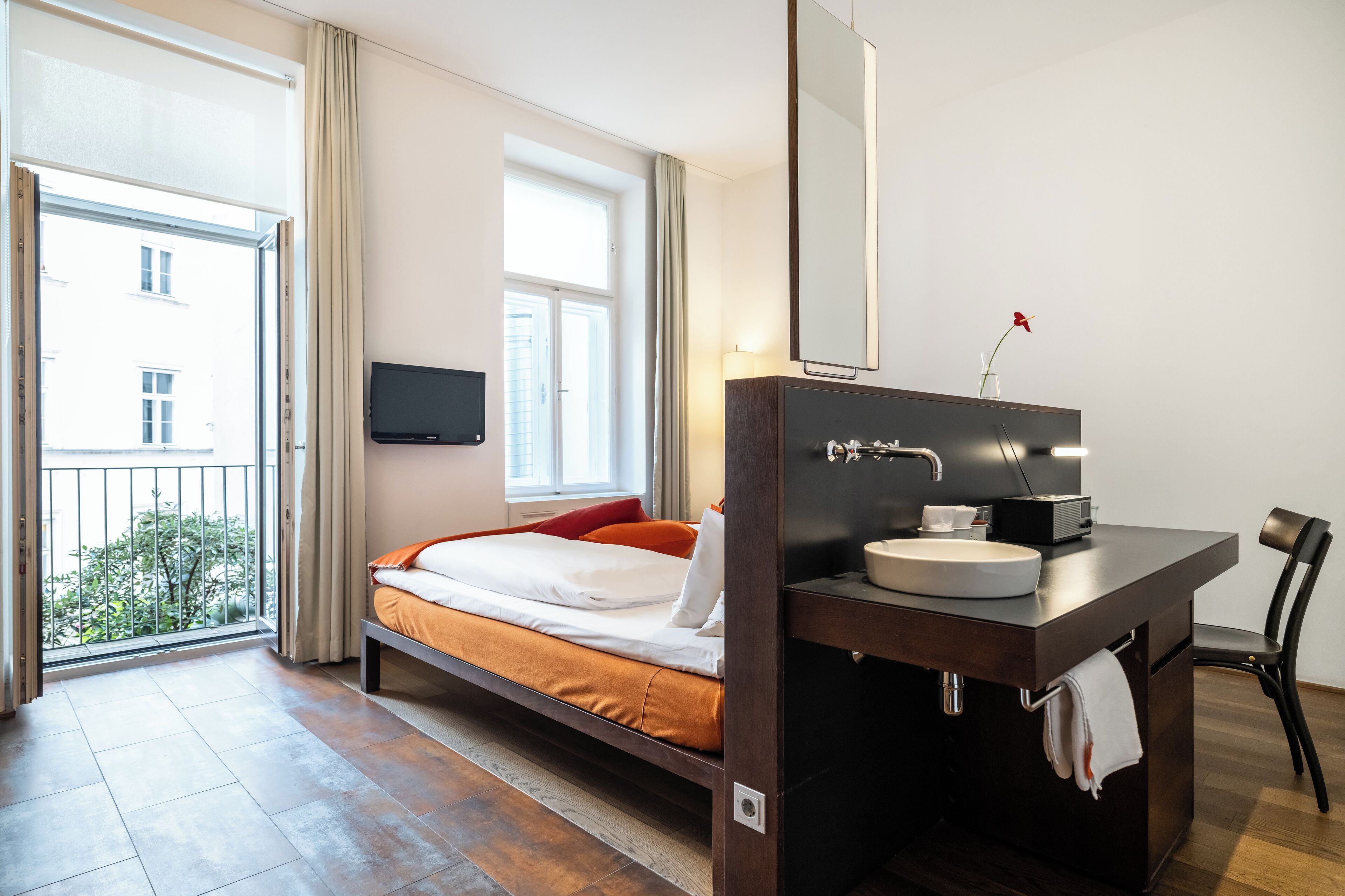 A hotel room with a bed, a bathroom with a sink and mirror, a TV on the wall, and a view of the outside.