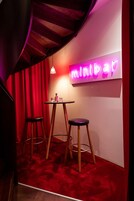 Uma área de bar com letreiro de neon, dois bancos e uma pequena mesa com garrafas.
