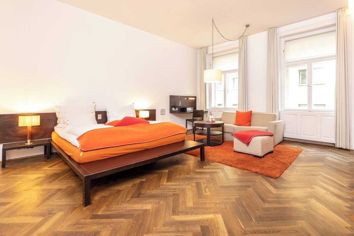 Una habitación de hotel con cama, mesitas de noche, televisor, sofá y un ventanal grande.