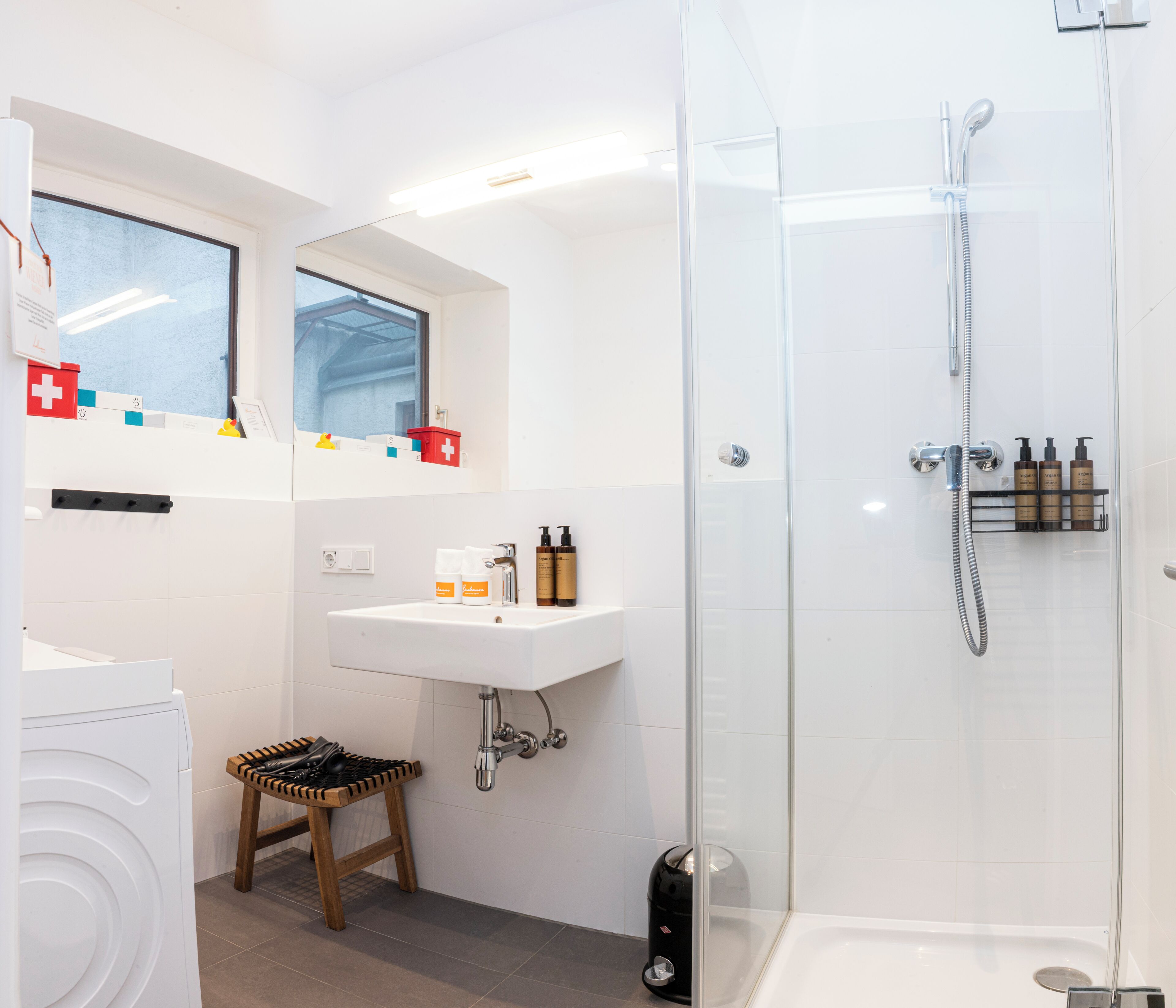 Un baño moderno con cabina de ducha de vidrio, un lavabo con artículos de aseo y un pequeño taburete.