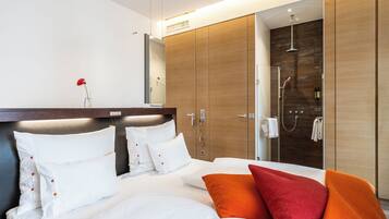 Una habitación de hotel moderna con una cama grande, un armario de madera, una zona de ducha y un pequeño jarrón con una flor roja sobre la mesita de noche.