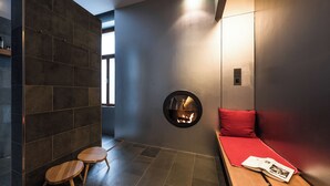 Un interior moderno con una pared de piedra, una chimenea circular, un banco con cojín rojo y una pequeña mesa de madera con una revista.