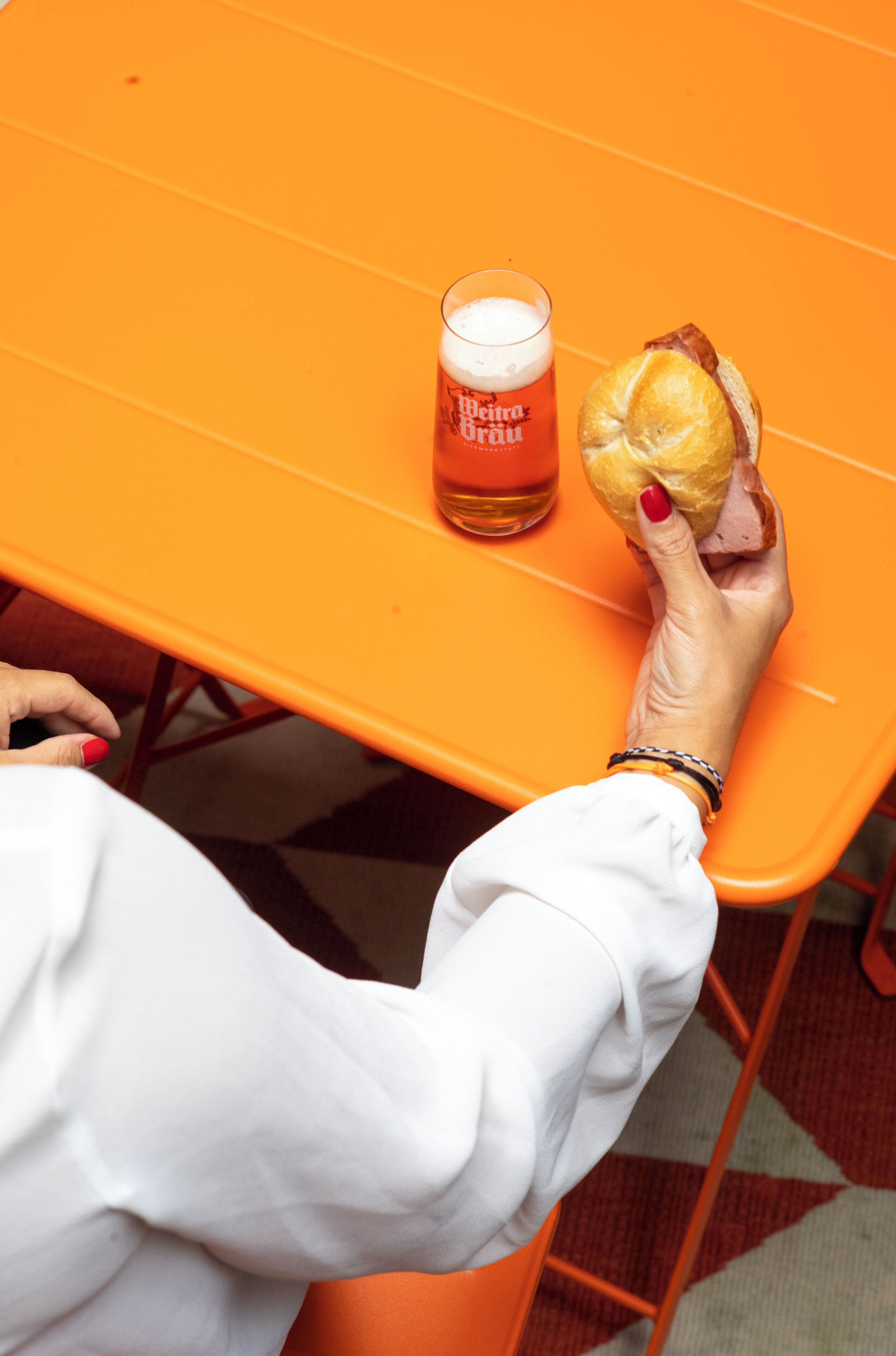 Una persona sosteniendo un sándwich y una jarra de cerveza sobre una mesa anaranjada.
