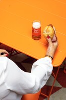 Uma pessoa segurando um sanduíche e um copo de cerveja sobre uma mesa laranja.
