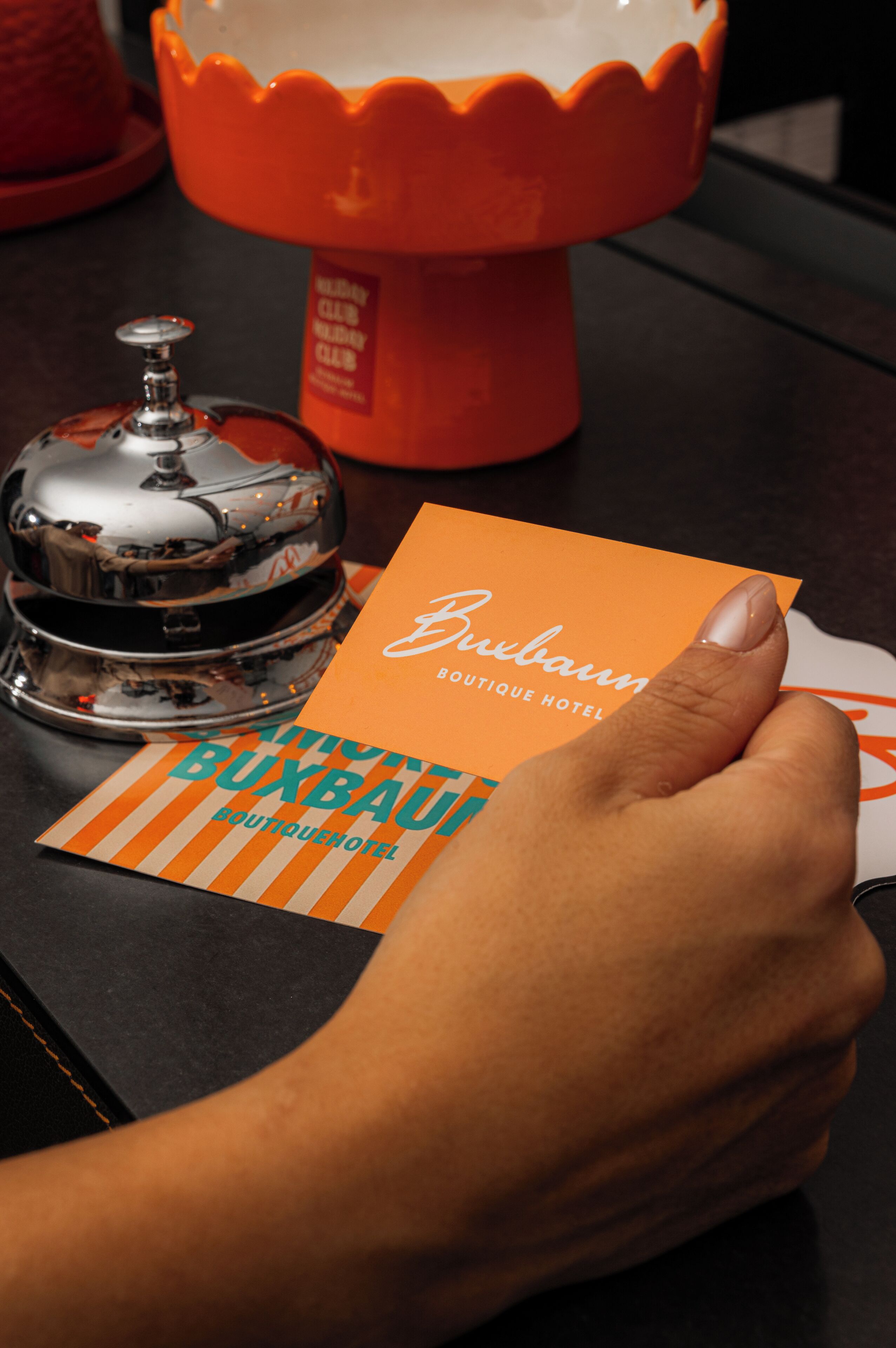 Una mano sosteniendo una tarjeta con la leyenda 'Buxbaum Boutique Hotel', una campana plateada y un candelabro rojo.