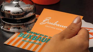 Una mano sosteniendo una tarjeta con la leyenda 'Buxbaum Boutique Hotel', una campana plateada y un candelabro rojo.