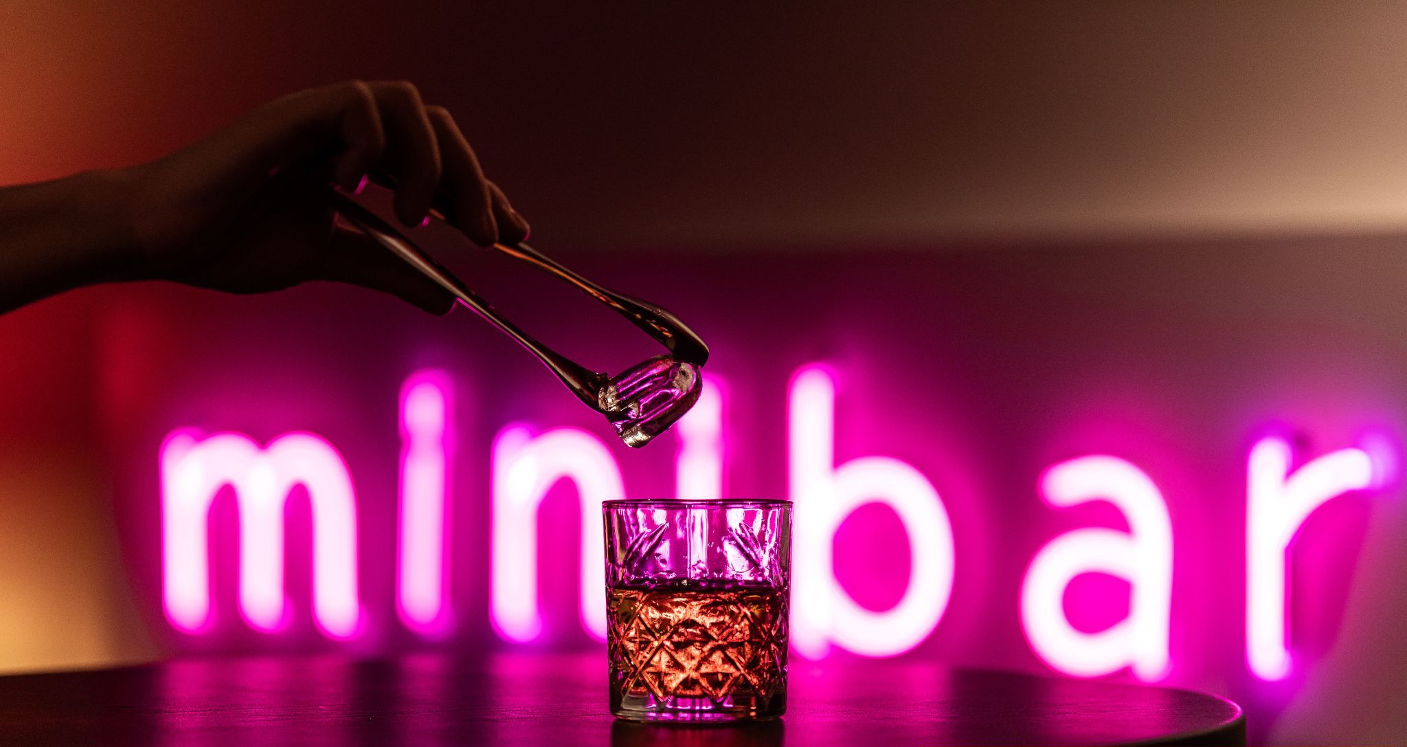 Une main tenant une pince au-dessus d’un verre au motif texturé, posé sur une table. Un panneau lumineux au néon est visible en arrière-plan.