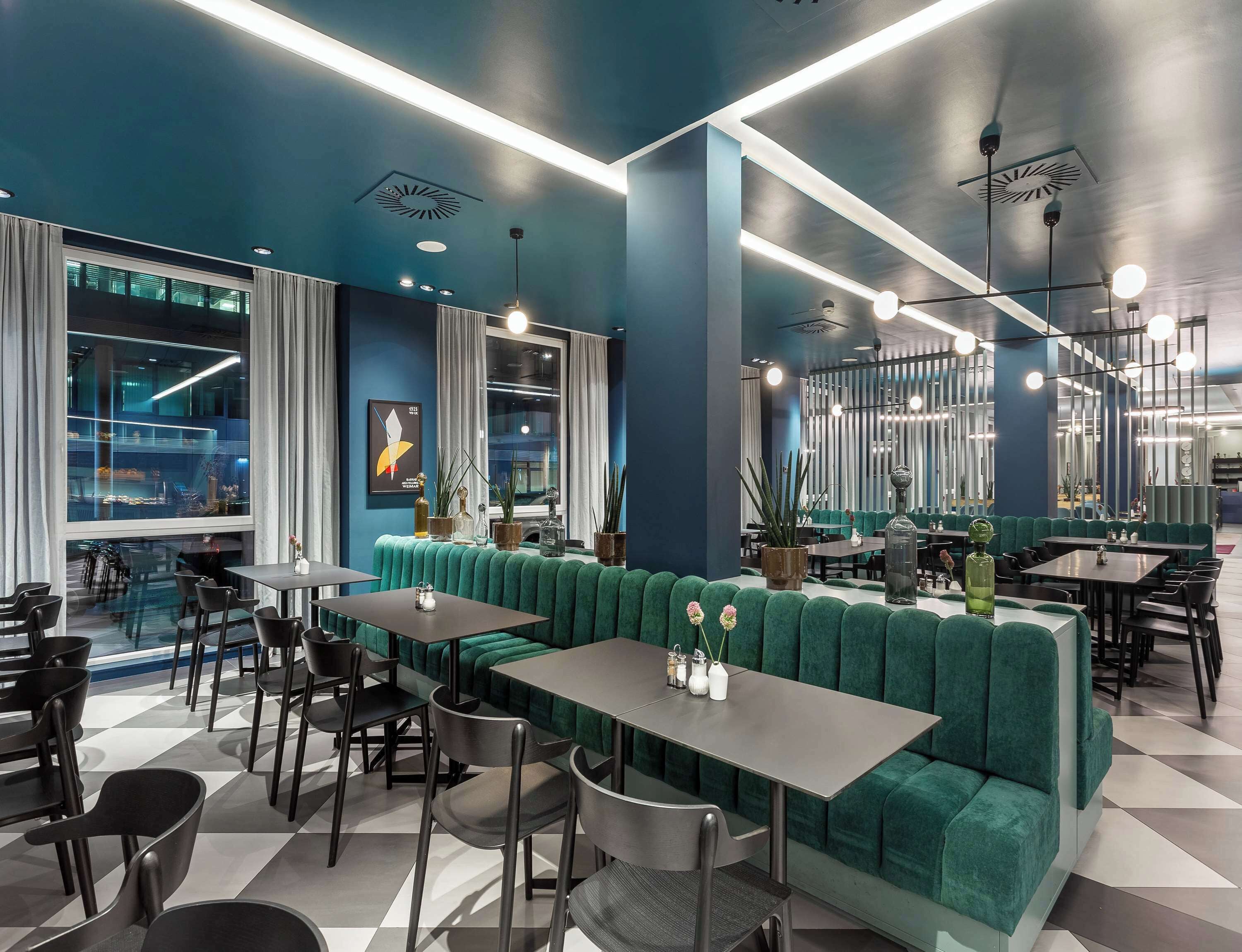 Un intérieur de restaurant moderne avec des banquettes recouvertes de tissu vert, des chaises noires et un sol à carreaux.