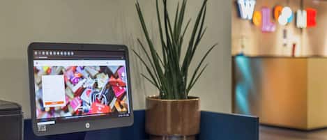 Una computadora de escritorio con monitor mostrando una página web, teclado, ratón, una planta en maceta y un mostrador de recepción con un letrero.
