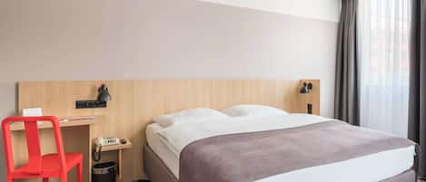 Habitación de hotel con cama, escritorio, silla, lámpara y un cubo de basura.