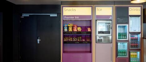 Un área de máquinas expendedoras con secciones identificadas para aperitivos, hielo y bebidas, que incluyen marcas como Premier Inn y Red Bull.