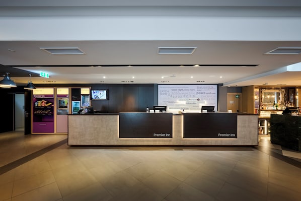 Una recepción de hotel con una pantalla digital mostrando un mensaje, una máquina expendedora y una zona de bar.