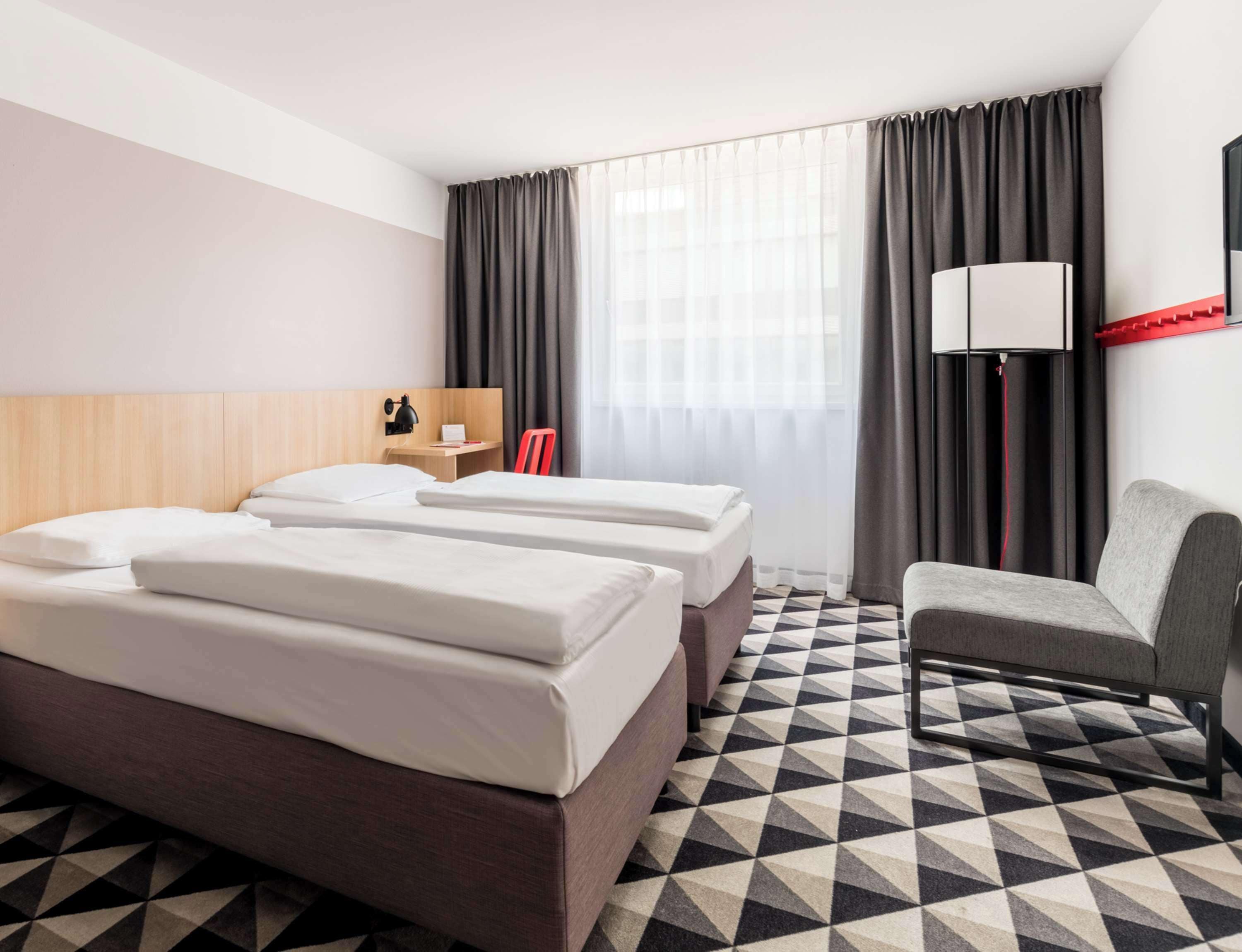 A hotel room with two beds, a desk, a chair, a lamp, and a large window with curtains.